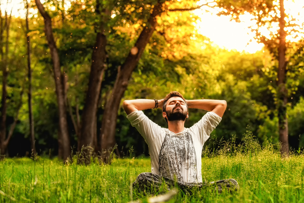A man sitting on grass in the park and stretching with eyes closed and hands behind head. Concept of meditation, dreaming, wellbeing and healthy lifestyle